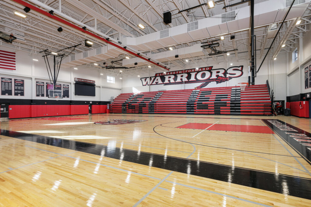 An indoor basketball court with red and black bleachers, featuring the “Elizabeth Forward Warriors” logo on the wall.