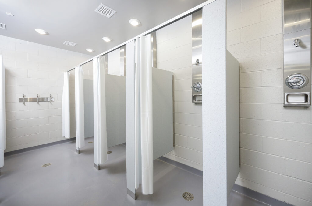 A row of public shower stalls with white curtains, gray partitions, and wall-mounted shower fixtures.