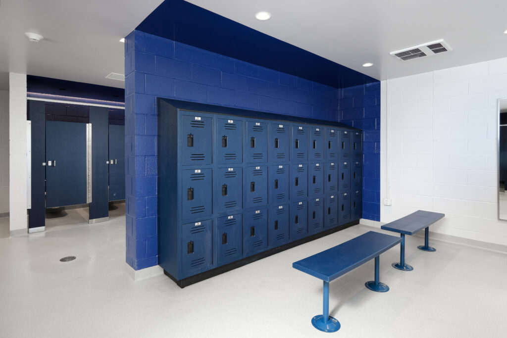 A locker room with rows of blue metal lockers, two blue benches, and blue-painted cinder block walls.