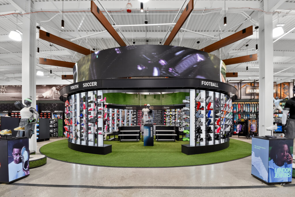 A circular sports store display for youth soccer and football cleats, featuring rows of footwear on curved shelving and a digital screen overhead.