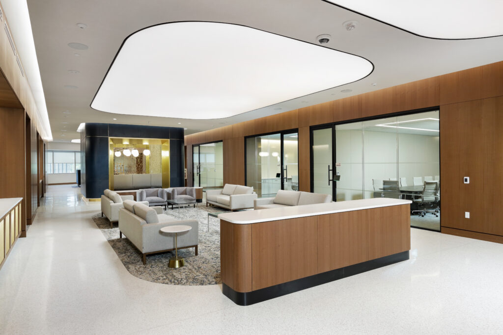 A modern office reception area with a wood-paneled front desk, a seating area with sofas on a patterned rug, and glass-walled conference rooms.