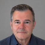Headshot of a middle-aged man with short, graying hair wearing a blue striped button-down shirt against a gray background.