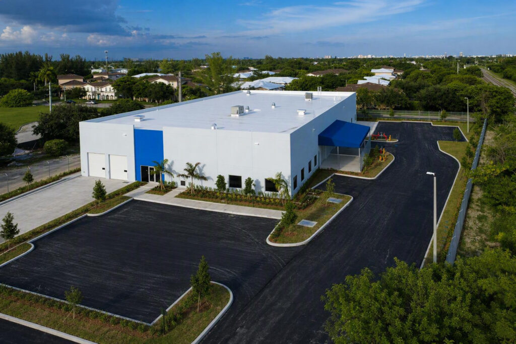 Aerial view of a white commercial warehouse building with blue accents, surrounded by a paved parking lot and green landscaping in a suburban area.