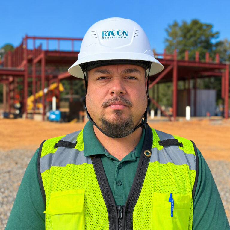 A man in a green polo shirt and high-visibility safety vest wearing a Rycon Construction hard hat, standing in front of a building under construction.