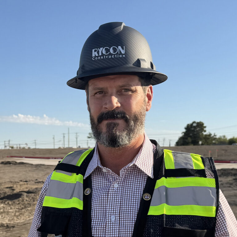A man wearing a Rycon Construction hard hat and a high-visibility safety vest at a construction site.